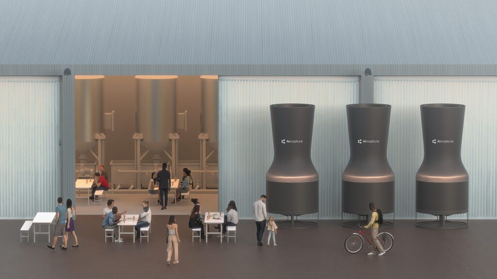 On the left — groups of people are seated at tables, with brewery equipment seen in the background in a warehouse. Three of AIrcapture's direct air capture devices are on the right side in front of the warehouse.